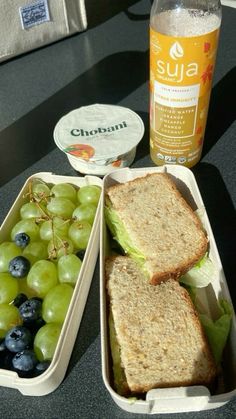 two plastic containers filled with sandwiches and grapes next to a bottle of soda on a table