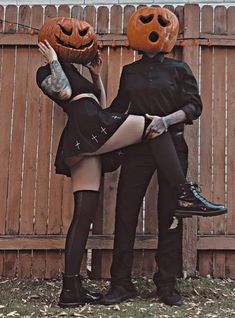 a man and woman dressed up in halloween costumes with pumpkins on their heads, standing next to a fence