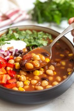 a spoon full of beans and vegetables being held up by someone's hand over the bowl