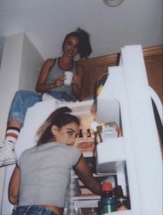 two young women sitting on top of an open refrigerator in a room with white walls