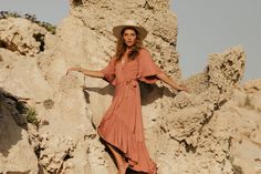 a woman in a dress and hat standing on rocks with her arms spread out to the side