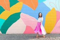 a woman standing in front of a colorful wall with flowers painted on it and wearing a pink skirt