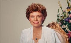 a woman sitting in front of a golden chair with flowers on the table behind her