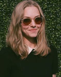 a woman with long blonde hair and sunglasses standing in front of a green hedge wall