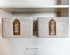 two canvas storage bins with tags on them in a kitchen cabinet, one is white and the other is brown