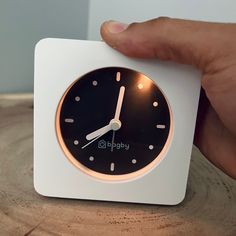 a hand holding a clock on top of a wooden table next to a white square