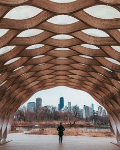 a person standing under a wooden structure in the middle of a park with buildings in the background