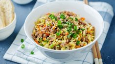 a white bowl filled with rice and vegetables next to chopsticks on a blue surface