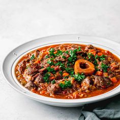 a bowl of stew with meat and vegetables on a white plate next to a napkin