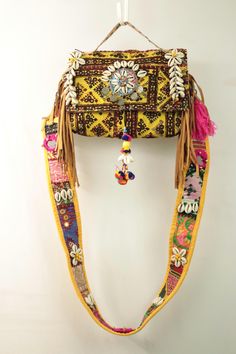 This handmade bag is a beautiful example of intricate craftsmanship. It features a broad strap decorated with white floral patterns, small round shells, and dark beads or coins. The body of the bag showcases detailed embroidery in earthy tones, with rows of pink tassels along the lower edge. The central part of the bag is adorned with large pink tassels, smaller multicolored pom-poms, and mirrored accents surrounded by embroidered designs. Additional shell decorations are symmetrically arranged Shell Decorations, Shell Bag, Large Bracelet, Jaune Orange, Pink Tassel, Boho Bag, Embroidered Design, Earthy Tones, Pom Poms