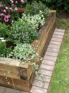 a wooden planter filled with lots of flowers on top of a brick walkway next to grass