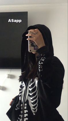 a woman wearing a skeleton hoodie taking a selfie with her cell phone while standing in front of a wall