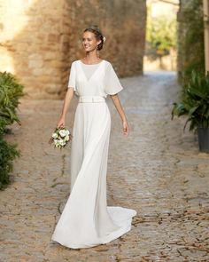 a woman in a white wedding dress walking down a cobblestone road holding a bouquet