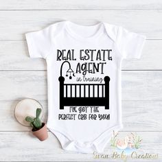 a white baby bodysuit with the words real estate agent in black on it, next to a potted plant