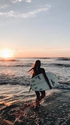 a woman walking into the ocean with a surfboard in her hand and sunset behind her