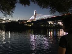 a woman standing on the side of a river next to a bridge with lights on it