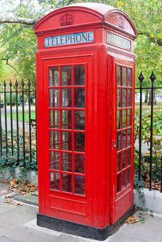 a red phone booth sitting on the side of a road next to a fence and trees