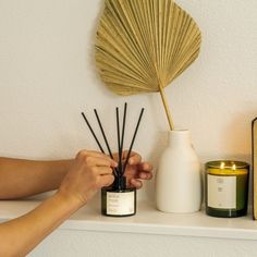 a person holding a candle next to a vase with reeds in it on a shelf