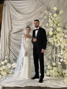 a bride and groom pose for a photo in front of a backdrop with white flowers