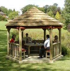 three people sitting at a table under a straw roofed gazebo in the middle of a field