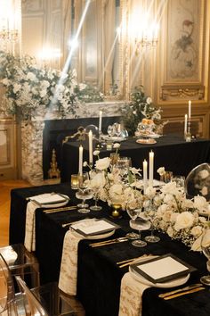 a dining table set up with white flowers and candles in front of an ornate fireplace