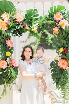 a woman holding a baby in her arms under a floral arch with pink and yellow flowers