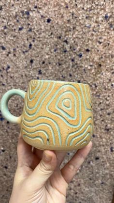 a hand holding a yellow and blue coffee mug in front of a carpeted wall