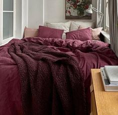 a bed with red sheets and pillows next to a window in a room that has white walls