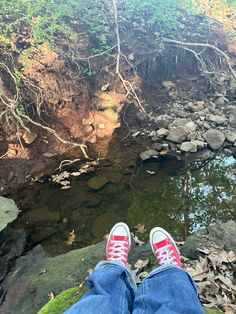 someone's feet with red and white sneakers standing in front of a stream surrounded by rocks