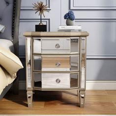 a white and gold mirrored night stand with two drawers on each side, next to a bed