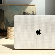 an apple laptop sitting on top of a wooden table next to a basket and painting