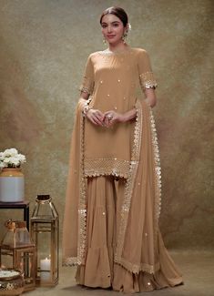 This exquisite Beige Georgette Kurta Sharara Set is a perfect blend of elegance and tradition. Made from georgette fabric, the beige kurta is adorned with delicate embroidery featuring mirrors, gota, sequins, and pearls, creating a stunning visual appeal. The neckline, hem, and sleeves are intricately embellished, complemented by small floral buttis that add a touch of grace. Teamed with a tiered sharara that showcases scattered mirror work, this ensemble exudes sophistication. The look is completed with a matching net dupatta, beautifully bordered and dotted with buttis, making it an ideal choice for Home Pooja, family gathering or festive occasions. Composition : Kurta and Sharara - Viscose Georgette and Dupatta - Soft net Care: Dry Clean Only and Vacuum Storage This product can be custo Latest Sharara Designs Party Wear, Golden Sharara, Beige Sharara, Mirror Work Sharara, Mirror Work Kurta, Bridal Crop Top, Beige Kurta, Kaftan Kurta, Georgette Kurta