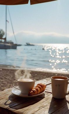 coffee and croissants sit on a wooden table at the beach with boats in the background