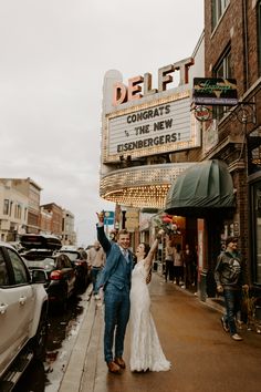 a newly married couple walking down the street in front of a movie theater holding their hands up