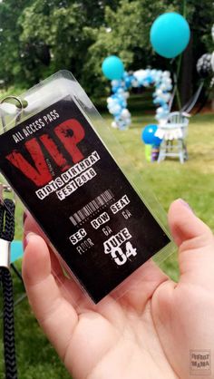 a person holding up a ticket for a concert in the park with balloons and streamers behind them