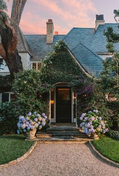 the front entrance to a house with flowers in pots on the driveway and trees around it