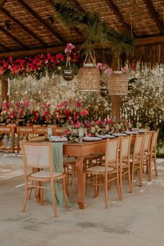 a wooden table topped with lots of chairs next to pink and white flowers on top of a lush green field