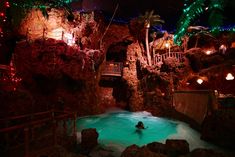 a person in a pool surrounded by trees and rocks with lights on the side of it