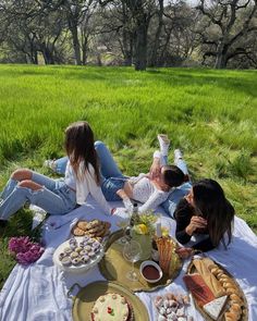 three women sitting on a blanket in the grass eating desserts and drinking tea together