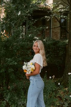 a woman standing in front of some trees holding flowers