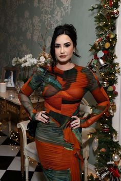 a woman standing in front of a christmas tree wearing an orange and green dress with long sleeves