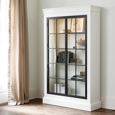 a white bookcase with glass doors in front of a window next to a wooden floor