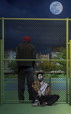 a man sitting on the ground in front of a fence with another man standing next to him