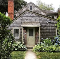 a gray house with a green door surrounded by bushes and flowers in front of it