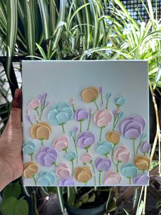 a person holding up a card with flowers on it in front of some potted plants