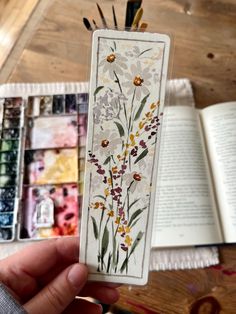 a person holding up a bookmark with flowers on it and paintbrushes in the background