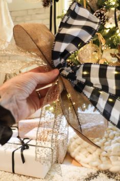 a hand holding a ribbon over a present box with christmas decorations around it and a tree in the background