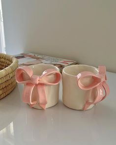 two mugs with pink bows on them are sitting next to a basket and magazine