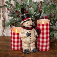 a snowman candle holder sitting on top of a wooden table next to two red and white checkered candles