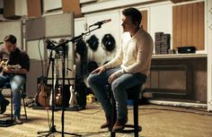 two men sitting on stools in front of microphones and recording equipment, with one playing the guitar